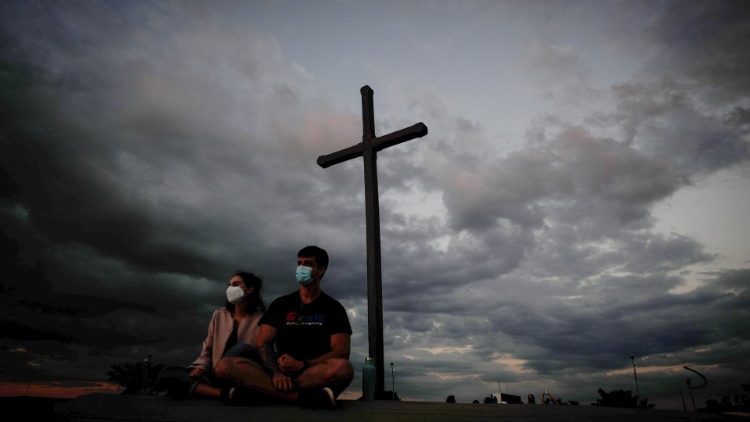 500-mil-mortes-covid-dom-orani Casal usando máscaras em frente a uma cruz. No fundo, um céu escuro e com muitas nuvens.