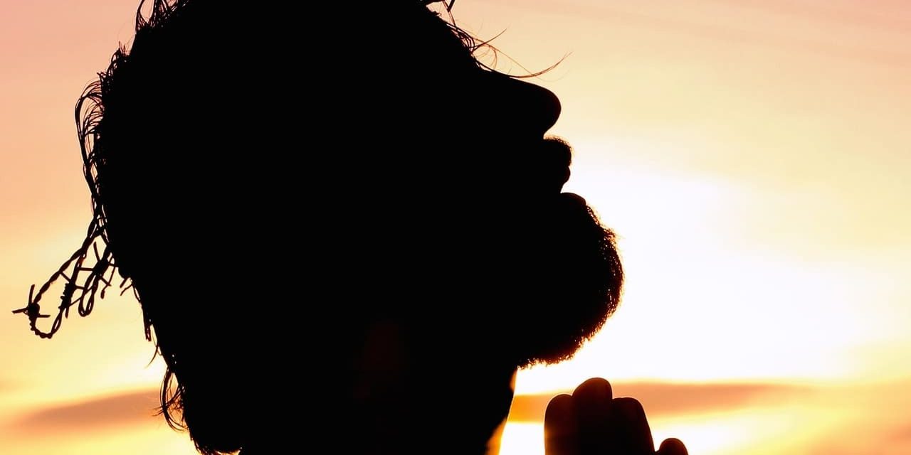 Silhueta de Jesus usando a coroa de espinhos enquanto ora; seu rosto está direcionado para o céu e há um pôr do sol ao fundo.