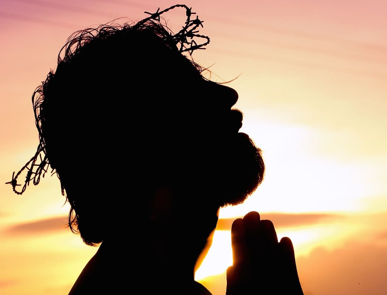 jesus-cristo Silhueta de Jesus usando a coroa de espinhos enquanto ora; seu rosto está direcionado para o céu e há um pôr do sol ao fundo.