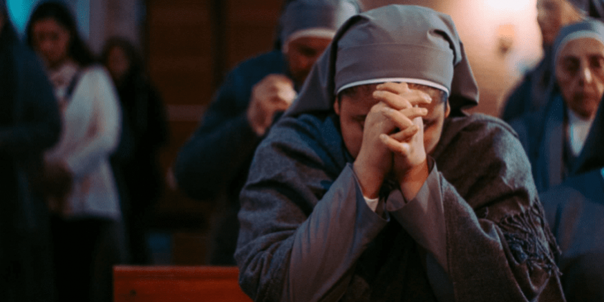 Mulher religiosa usando hábito cinza e rezando com as mãos juntas e a cabeça baixa