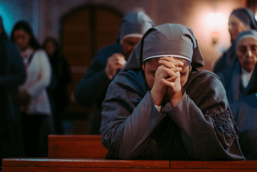 Mulher religiosa usando hábito cinza e rezando com as mãos juntas e a cabeça baixa