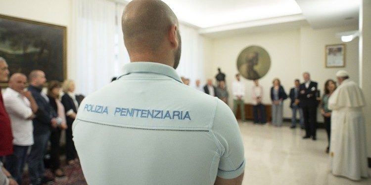 papa-detentos-roma Policial italiano visto de costas; sua camiseta diz "Polizia Penitenziaria". É possível ver o Papa Francisco conversando com uma pessoa no canto direito.