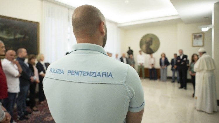 papa-detentos-roma Policial italiano visto de costas; sua camiseta diz "Polizia Penitenziaria". É possível ver o Papa Francisco conversando com uma pessoa no canto direito.