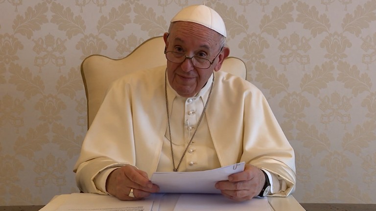 Papa Francisco segura papel sobre mesa de madeira enquanto olha para a câmera.